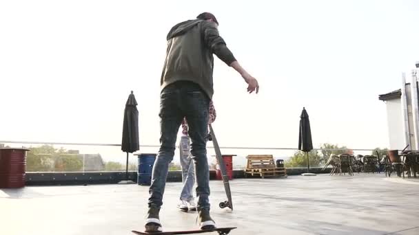 Bonito rapaz e menina estão se divertindo com skates no telhado de um edifício industrial. Skateboarder monta com sua namorada no telhado do edifício. Ao ar livre — Vídeo de Stock
