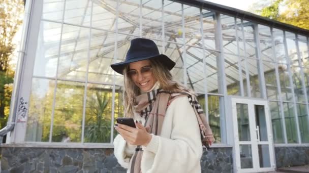 Молодая красивая женщина в шляпе болтает с другом, используя смартфон в городском парке. Портретный снимок . — стоковое видео