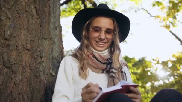Nära upp skott av ung kvinna i snygg hatt skriva något i anteckningsboken samtidigt slappnar av i stadsparken. — Stockvideo