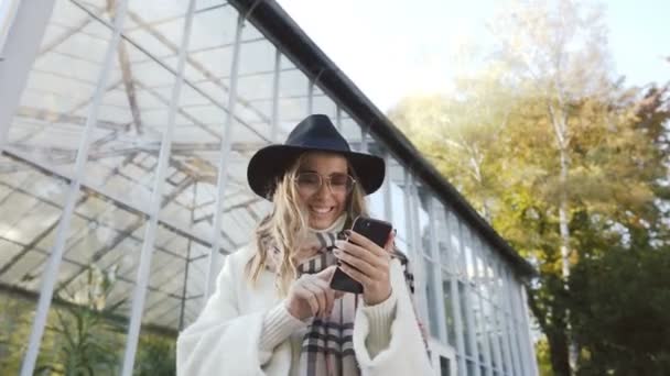 Mujer de pelo rubio feliz en gafas de sol escribiendo mensaje en el teléfono inteligente en el fondo del parque . — Vídeo de stock