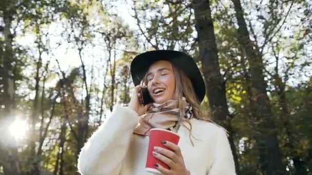 Porträt einer schönen Frau, die Kaffee trinkt und mit jemandem auf ihrem Smartphone spricht, während sie im Park spaziert. — Stockvideo