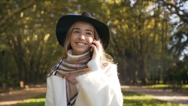 帽子とスカーフ秋の公園で歩いている間彼女のスマート フォンを使用して誰かと話している幸せなスタイリッシュな女の子のショットを閉じる ドーリー ショット — ストック動画