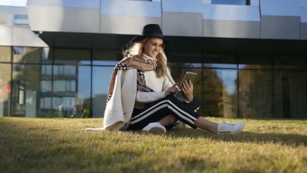 Joven chica sonriente relajándose en la hierba verde cerca del edificio moderno mientras usa su tableta . — Vídeos de Stock