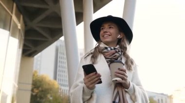 Fularlı kahve fincan ile kulaklık ile yürüme ve akıllı telefon kullanarak sohbet şirin kız.
