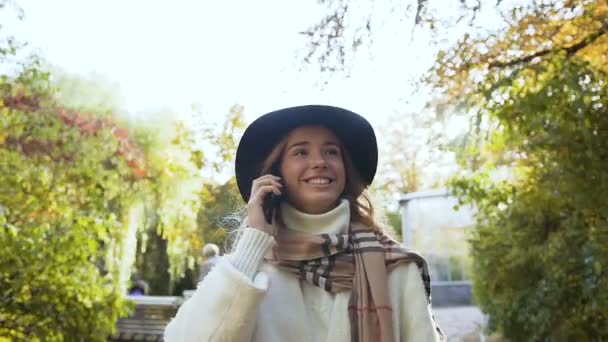 时尚的金发女孩戴帽子 白色外套微笑着 在绿色公园散步时 用手机与人交谈 阳光灯 — 图库视频影像