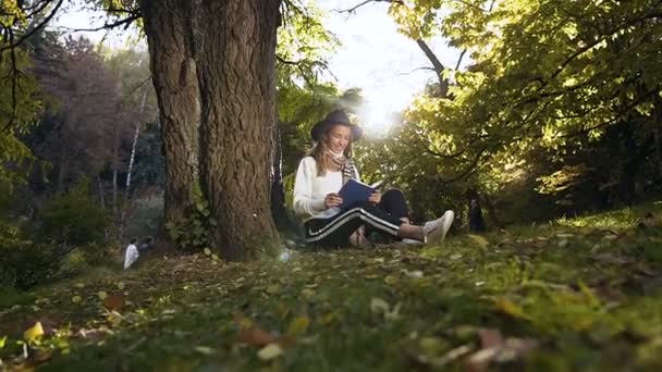 Snygg tjej i hatten avkopplande i parken, läsa bok medan du sitter på gräset. — Stockvideo