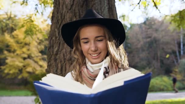 Mulher de cabelo loira sorridente em chapéu elegante e cachecol no parque da cidade livro de leitura . — Vídeo de Stock