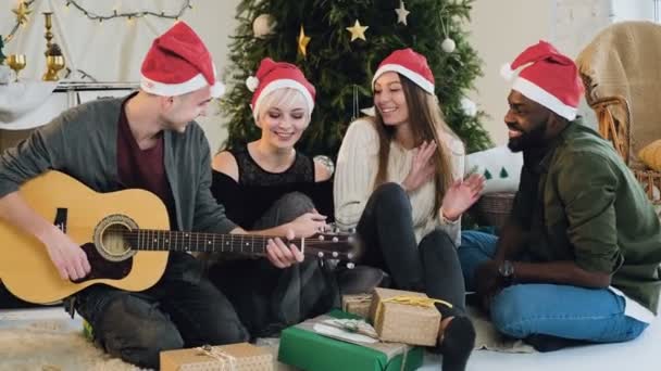 Un tipo vestido con sombrero de Santa Claus toca una guitarra cerca del árbol de Navidad. Los amigos celebran la Navidad en la habitación del loft, bailan cantan y se divierten . — Vídeo de stock