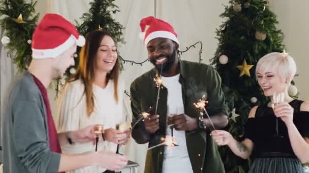 A cég fiatal szép különböző fajú emberek is szórakoztató táncol tovább, hogy a karácsonyfa. Meg ünnepelni karácsony és ital a bor, és éget a bengáli fények — Stock videók