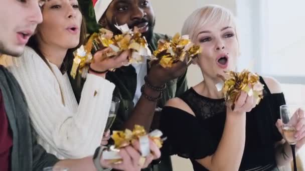 Крупным планом. Друзья разных рас выдувают золотые конфетти из рук в рождественской шляпе. Рождество и Новый год. Slow motion 4k shot — стоковое видео
