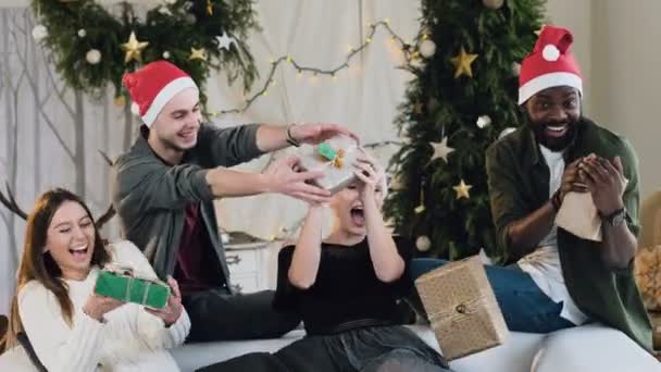 Amigos ativos e sorridentes pegar os presentes. presentes voar em suas mãos. mulheres jovens e homens em trajes de Ano Novo celebram o Natal no fundo das decorações de Natal com árvore de Natal — Vídeo de Stock