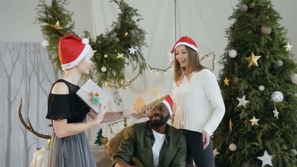 Young couple of cheerful people in santa claus hats cheerfully presents gifts to their friends on the background of a Christmas tree. Concept of celebrate New Years, Christmas or birthday party — Stock Video