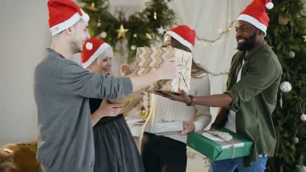 Young cheerful friends in Santa hats celebrating Christmas are standing before christmas tree, holding present boxes and having fun. Concept of celebrating New Year, Christmas and winter holidays — Stock Video