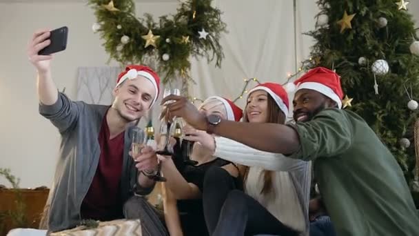 Ευτυχής παιδιά και κορίτσια ντυμένοι με καπέλα Άγιου Βασίλη κρατώντας ποτήρια κρασί κάνει selfie φωτογραφία για το smartphone κοντά τις διακοσμήσεις Χριστουγέννων — Αρχείο Βίντεο