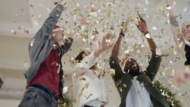 Jóvenes felices bailan lanzando confeti de oro en la fiesta de celebración. Hermosas chicas y chicos están celebrando el Año Nuevo o Navidad, que están brillando con un confeti brillante — Vídeo de stock