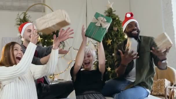 Felizes amigos sorridentes estão sentados no sofá pegar os presentes caixas. Presentes voam em suas mãos. Jovens mulheres e homens em trajes de Ano Novo celebram o Natal no fundo do Natal — Vídeo de Stock