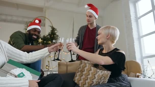 Amigos alegres bebiendo tostadas mientras celebran la Navidad o Año Nuevo, en un acogedor apartamento, sentados en el sofá antes del árbol de Navidad. grupo de jóvenes de raza mixta en la fiesta de Navidad con — Vídeos de Stock