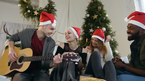 A group of cheerful young people having fun at home near a Christmas tree, one of the guys playing guitar — Stock Video