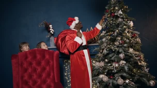 Papai Noel colocando brinquedo na árvore de natal, enquanto duas meninas espionando para ele . — Vídeo de Stock