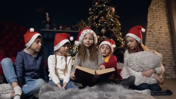 Κομψό χαριτωμένο παιδιά ανάγνωση του βιβλίου, χαλαρώνοντας σε ένα δωμάτιο κοντά στο χριστουγεννιάτικο δέντρο. — Αρχείο Βίντεο