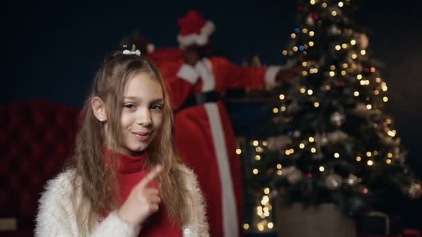 Маленька мила дівчинка кладе палець на губи, просячи бути тихіше, поки Санта Клаус кладе подарунок . — стокове відео