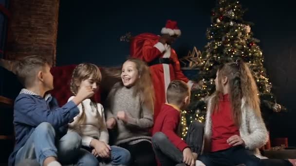 Fröhliche süße Kinder spielen, lachen und geben fünf auf dem Hintergrund mit afrikanischen Mann im Weihnachtsmann-Kostüm. — Stockvideo