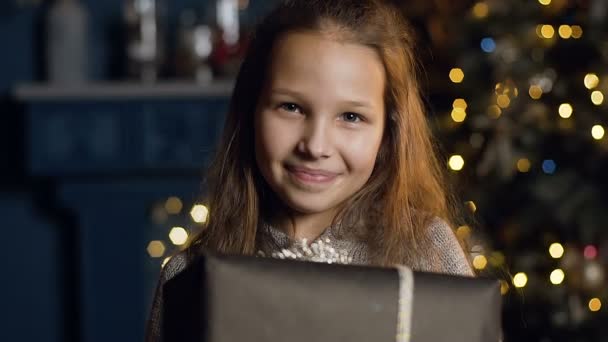 Primer plano de chica linda sosteniendo presente y mirando a la cámara en el fondo del árbol de Navidad . — Vídeos de Stock