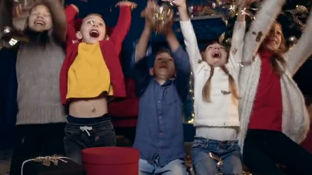 Retrato de niños felices sentados cerca del árbol de Navidad con los regalos y empezar a lanzar confeti . — Vídeo de stock