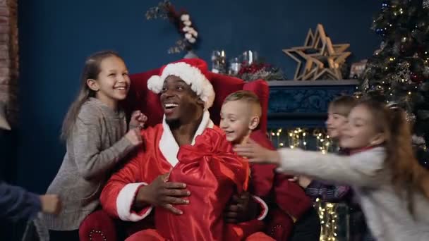 Санта Клаус в шляпе сидит на красном кресле, пока пятеро детей неожиданно бегут и обнимают его . — стоковое видео