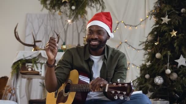 Usmívající se Afričan s kytarou a Bengálsko světlo sedí na podlaze v třpytivé zlaté konfety na pozadí vánoční stromeček. Koncept oslav nového roku, oslavy