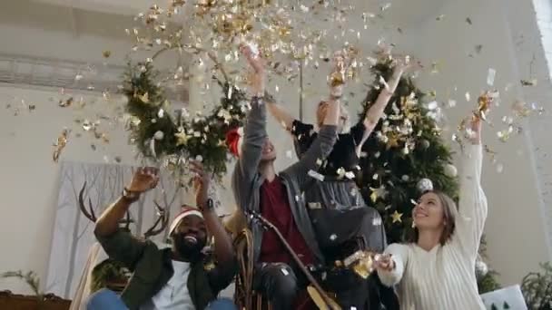 Two handsome young guys wearing in Santas hats with their girlfriends throw golden confetti at celebrating at the New Years or celebrating Christmas. Group of happy friends laughing on Christmas Eve — Stock Video