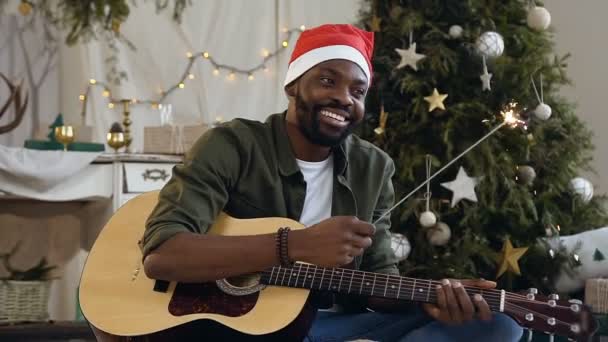 Guapo afro americano chico con guitarra y bengala luz feliz está sentado en el suelo en un brillante confeti dorado en el fondo de un árbol de Navidad y decoraciones de Navidad — Vídeos de Stock