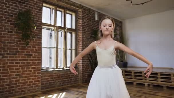 Young slim ballerina dressed in white tutu trains gracefully in pointe ballet shoes in ballroom. Graceful ballerina performs ballet positions in white tutu skirt near large window and red brick wall — Stock Video