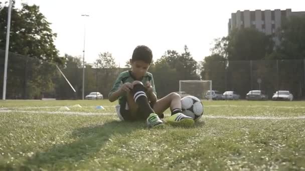 Menino sentado no campo de futebol depois de treinamento duro fixação leggings futebol . — Vídeo de Stock