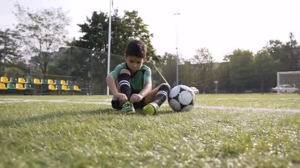 Menino consertando seus tênis enquanto descansa no campo de futebol após o campo de futebol duro . — Vídeo de Stock