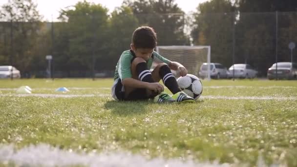Pequeño jugador de fútbol arreglando sus zapatillas de deporte mientras está sentado en el campo de fútbol . — Vídeo de stock