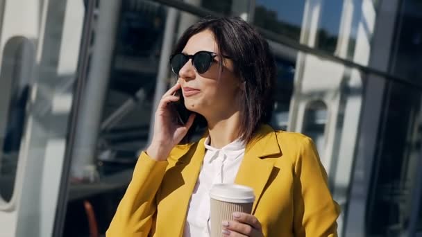 Porträtt av en vacker affärskvinna i kostym promenader nära businesscenter i staden, kommunicerar via smartphone och dricka kaffe. Begreppet nya affärer, kommunikation, bankir, manager. — Stockvideo