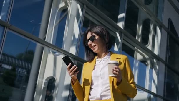 Sabah iyi bir ruh hali içinde takım elbise giymiş güzel genç iş kadını smartphone kullanarak ve kahve içme şehir sokaklarda işe gider — Stok video