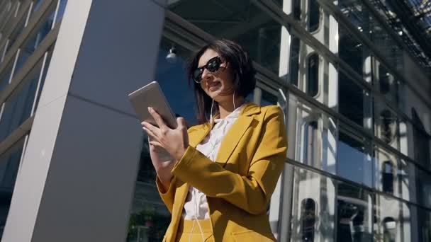 Retrato de mujer de negocios en traje sosteniendo en la mano y utiliza tableta. Mujer joven usando tableta pantalla táctil al aire libre, en el fondo moderno edificio de vidrio de oficina — Vídeo de stock