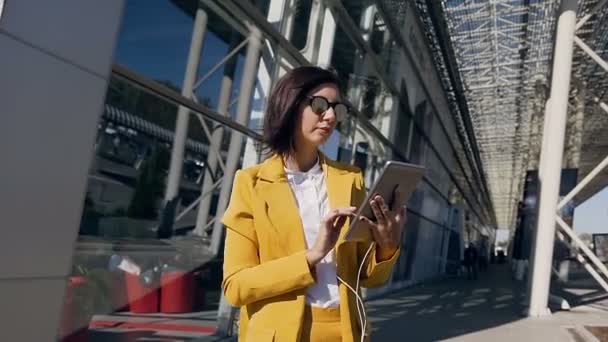 ビジネス センターの背景に都市の通りに立っているタブレット コンピューターを使用してヘッドフォンの若い美しい女性実業家 — ストック動画