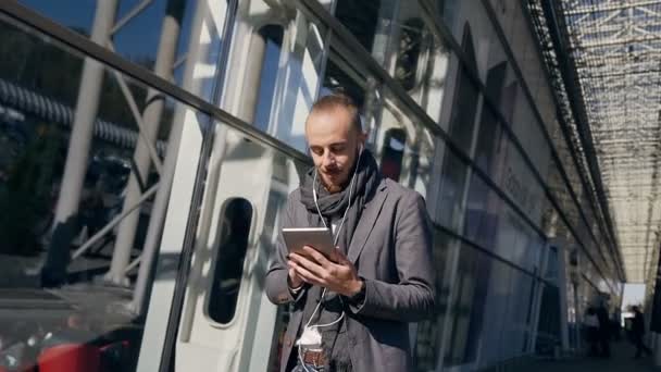 Glad kaukasiska affärsman använder surfplatta stående nära stora kontor centrum windows utomhus. Ung man i hörlurar rullning sidor, knacka på pekskärmen, kontrollera finansiella diagram på — Stockvideo