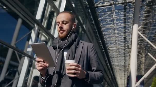 Hombre de negocios caucásico utilizando la aplicación de negocios en la computadora tableta, caminando en la ciudad durante una pausa para el almuerzo de trabajo. Guapo joven hombre de negocios que se comunica en la tableta, ir confiado, beber café — Vídeo de stock