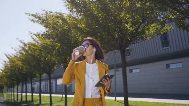 Joven mujer de negocios en traje y gafas de sol es mantiene una tableta en las manos y beber café en la calle urbana. Al aire libre — Vídeos de Stock