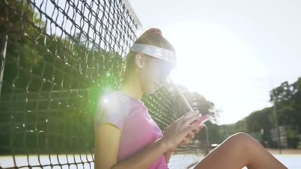 Menina do esporte sentado em um campo de ténis perto da rede e usando telefone inteligente . — Vídeo de Stock