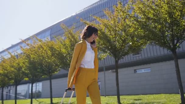 Elegante joven mujer de negocios en traje elegante tira de una maleta, hablando en su teléfono inteligente, se apresura a una reunión de negocios. Atractiva mujer de negocios que va en un viaje de negocios tirando de su maleta — Vídeos de Stock