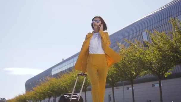 Ділова жінка в костюмі і сонцезахисних окулярах розмовляє по смартфону під час прогулянки з ручною поклажею біля вокзалу або аеропорту. Молода жінка щаслива, використовуючи мобільний додаток для розмови — стокове відео