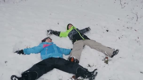 Two brothers in the colored winter clothes lay on the snow in forest and play waving with hands and feet. Winter holiday. December, January — Stock Video