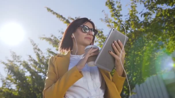 Mulher bonita em óculos de sol segurando café quente na xícara enquanto ela envia uma mensagem no computador tablet. Ao ar livre. Estudante do sexo feminino ou mulheres de negócios tablet computador e beber café perto — Vídeo de Stock