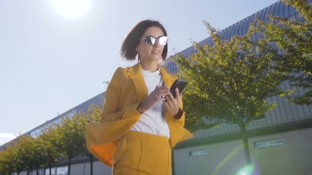 Retrato de una atractiva joven empresaria en gafas de sol caminando por la calle urbana y utilizando un teléfono inteligente moderno en el exterior. Joven mujer de negocios profesional usando smartphone para salir al aire libre — Vídeos de Stock