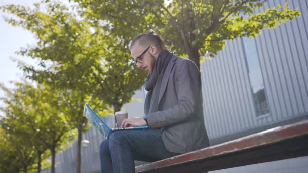 Przystojny biznesmen w okularach, ubrany w płaszcz z szalik za pomocą laptopa na przerwę na zewnątrz siedząc na ławce w parku, w pobliżu centrum biznesowego picie kawy — Wideo stockowe